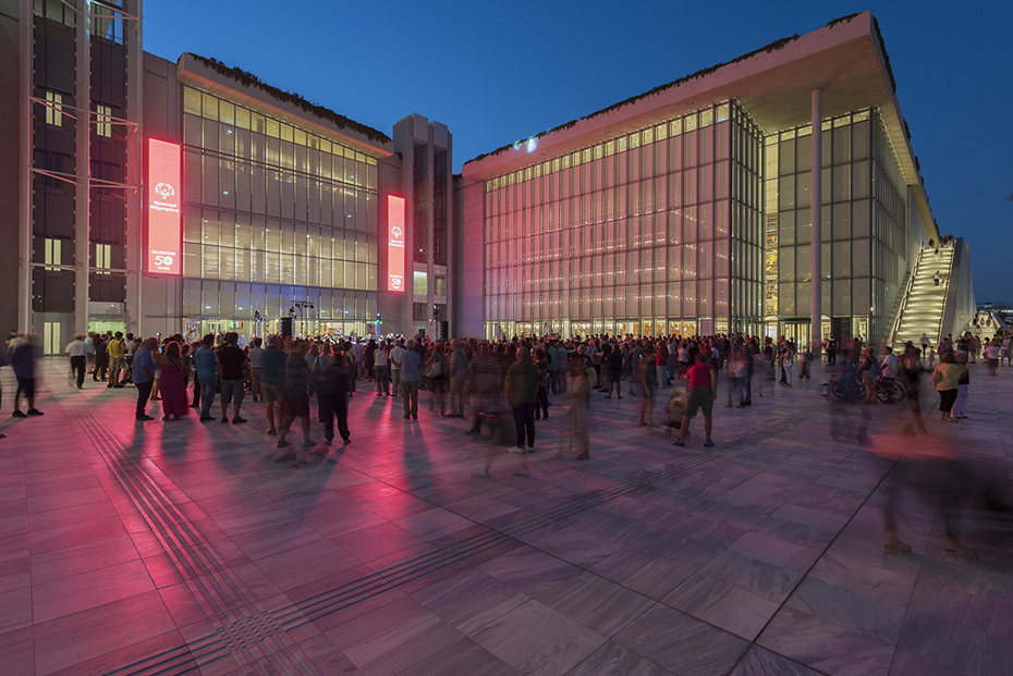Φωτογραφία που δείχνει πλήθος ανθρώπων να στέκονται όρθιοι ανάμεσα στον εξωτερικό χώρο που δημιουργούν δύο γυάλινα κτήρια