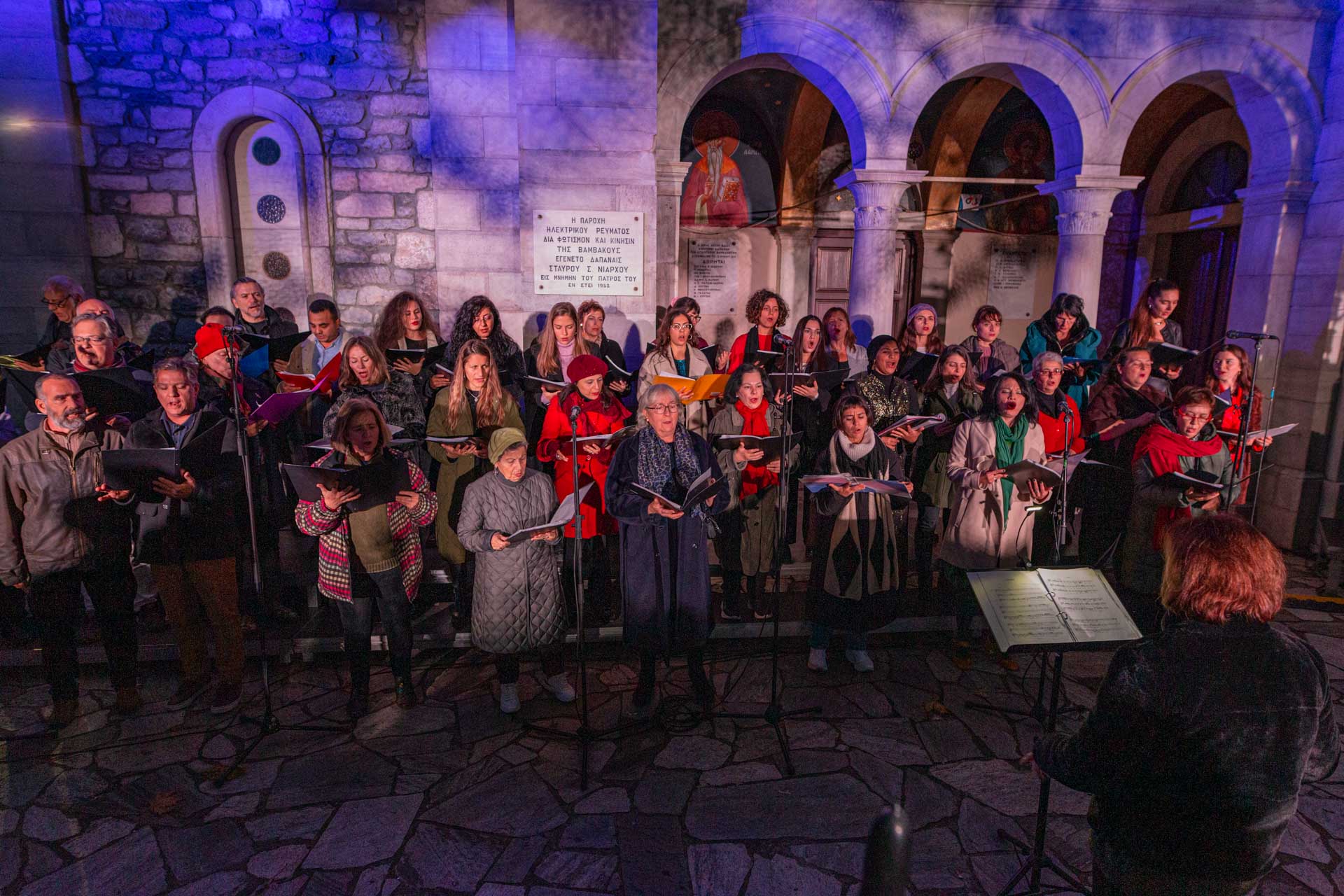 Η Διαπολιτισμική Χορωδία της Εθνικής Λυρικής Σκηνής στέκεται μπροστά από την εκκλησία του χωριού και τραγουδά ακολουθώντας τις οδηγίες της μαέστρου. 