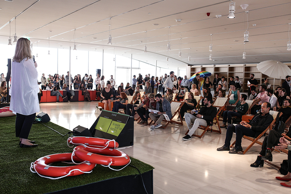 Μια γυναίκα δίνει ομιλία σε μια σκηνή με τεχνητό γρασίδι και μια μπάλα θαλάσσης, ενώ το κοινό κάθεται σε καρέκλες γκαζόν με ομπρέλες θαλάσσης απλωμένες γύρω από την αίθουσα.