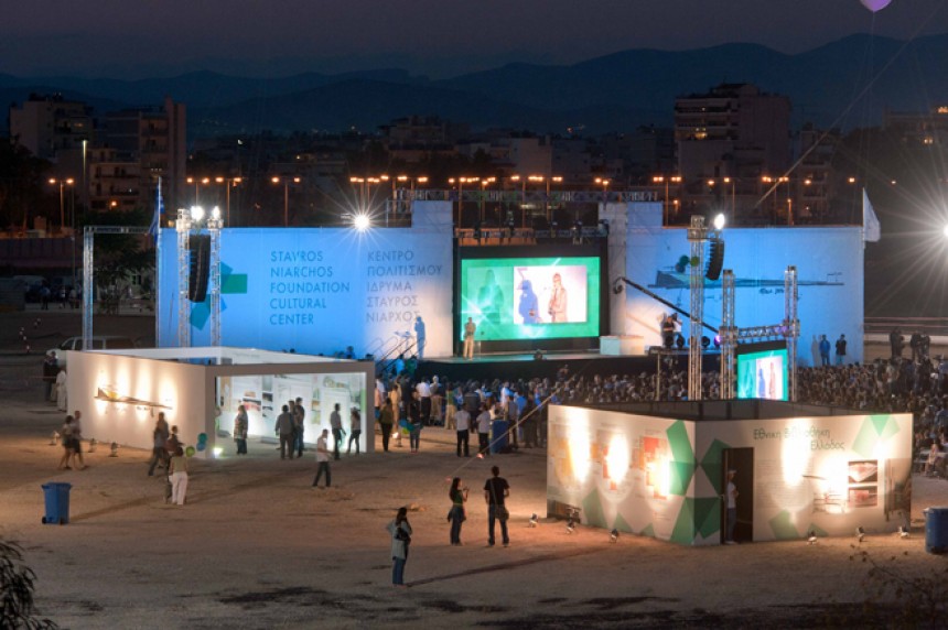 Μακρινή φωτογραφία τραβηγμένη τη νύχτα η οποία δείχνει πλήθος κόσμου σε ένα εργοτάξιο να κοιτούν προς τη σκηνή 