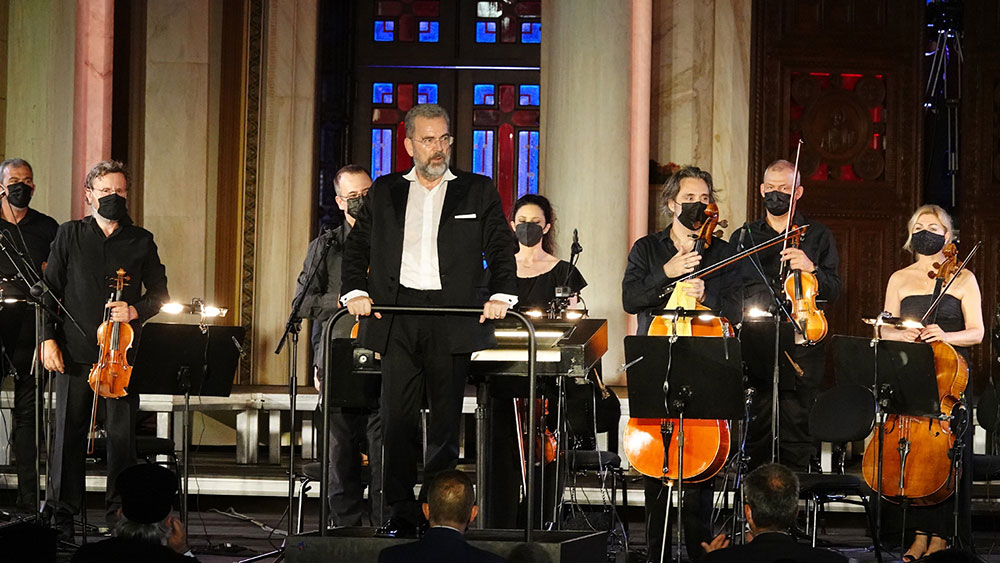 Πάνω σε μια σκηνή στέκεται όρθια μια ορχήστρα όπου τα μέλη της είναι ντυμένα στα μαύρα και μπροστά ο μαέστρος κοιτάει το κοινό