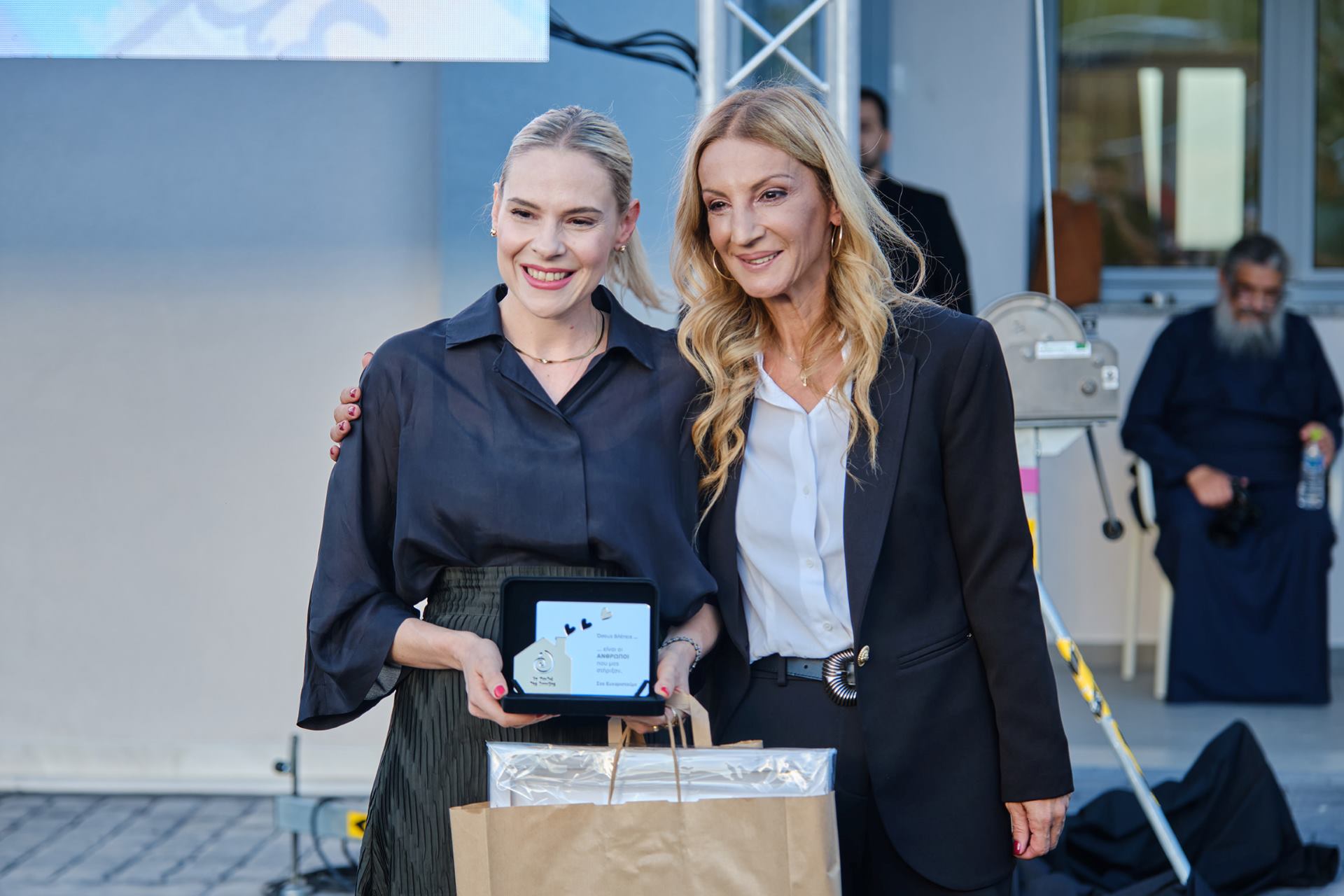 Δύο γυναίκες στέκονται δίπλα-δίπλα και χαμογελούν, η μία κρατάει μια τιμητική πλακέτα και μια τσάντα. 