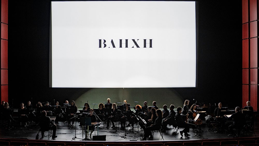 Φωτογραφία από την κεντρική σκηνή της Εθνικής Λυρικής Σκηνής στην οποία βρίσκεται όλη η ορχήστρα και από πίσω προβάλεται το όνομα του έργου ΒΛΗΧΗ 