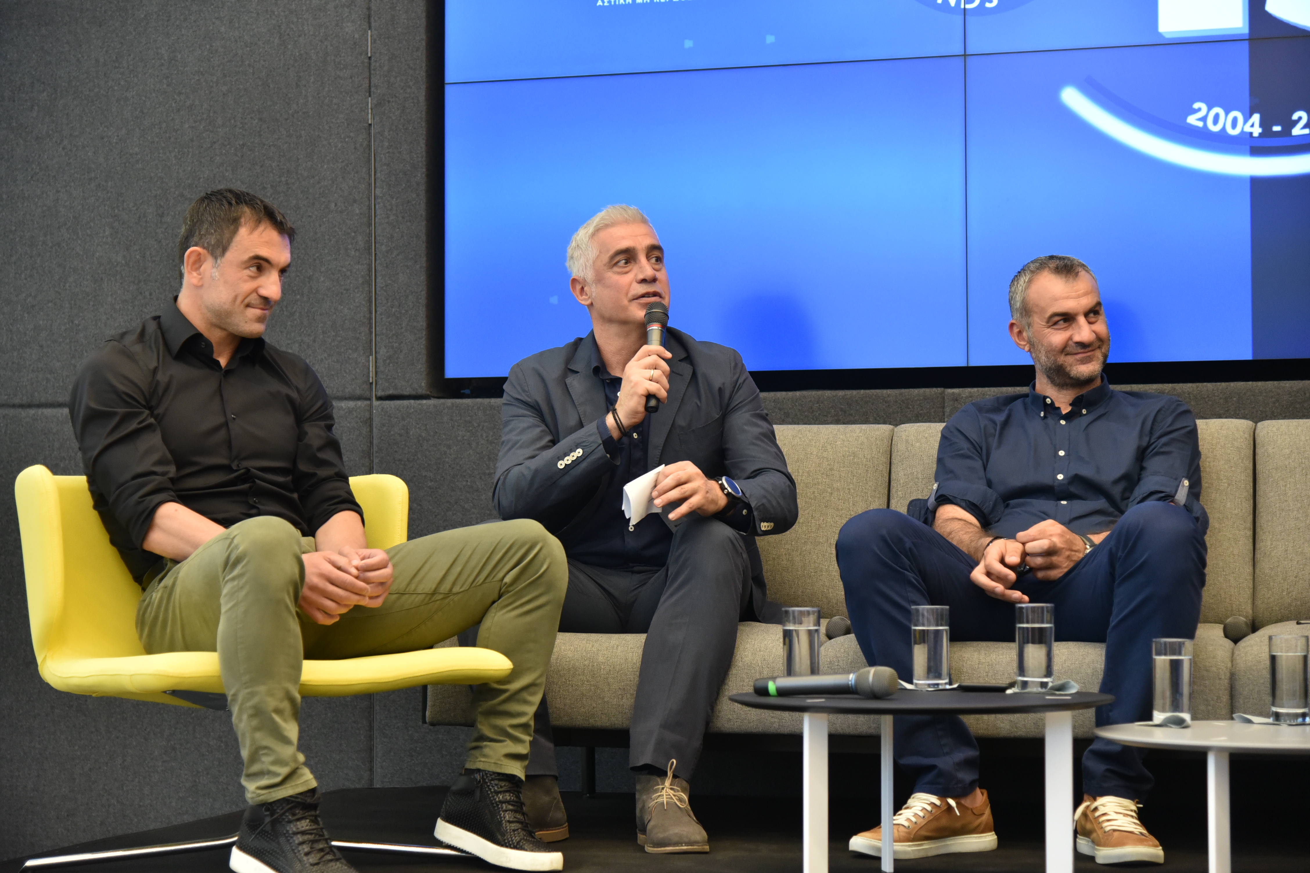 Κοντινή φωτογραφία τριών αντρών οι οποίοι κάθονται και συζητούν και ο ένας κρατάει ένα μικρόφωνο στα χέρια του και μιλάει 