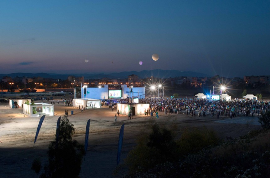 Μακρινή φωτογραφία τραβηγμένη τη νύχτα η οποία δείχνει πλήθος κόσμου σε ένα εργοτάξιο να κοιτούν προς τη σκηνή 