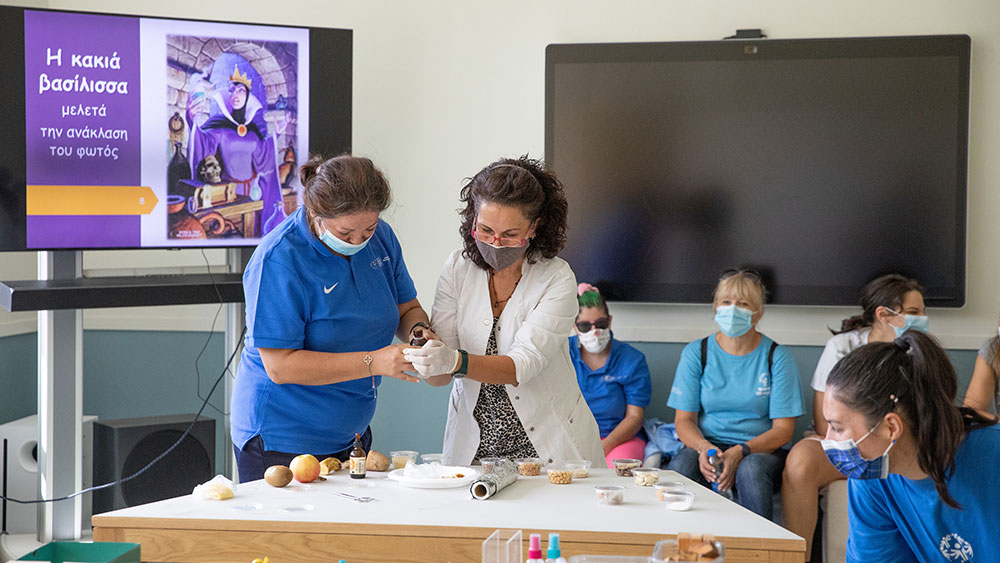 Δύο γυναίκες, η μία από τις οποίες φοράει εργαστηριακή ρόμπα, κάνουν επίδειξη πάνω από ένα τραπέζι με διάφορα συστατικά τροφίμων.