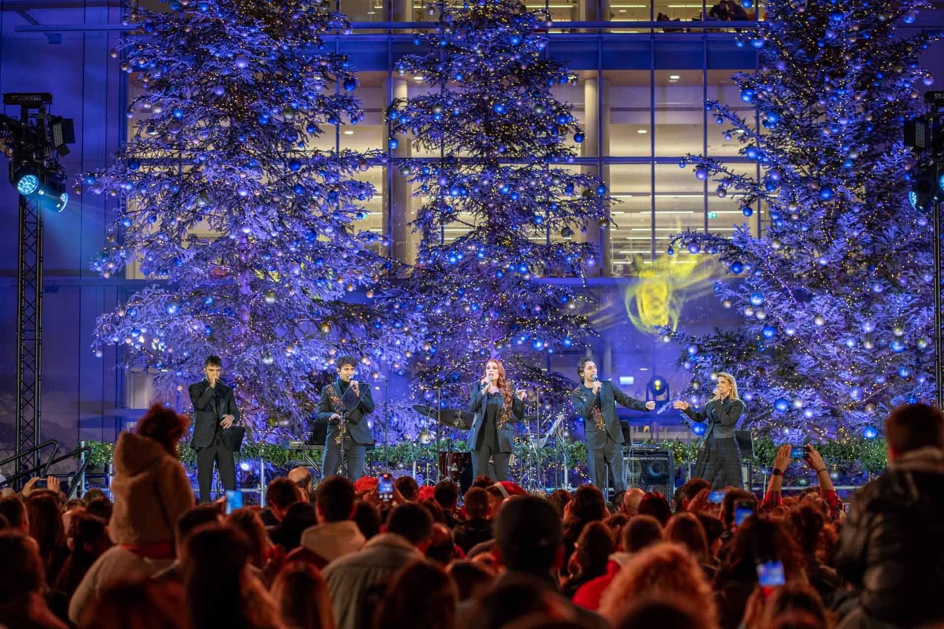 Μια ομάδα μουσικών στη σκηνή, με μεγάλα χριστουγεννιάτικα δέντρα στο φόντο.