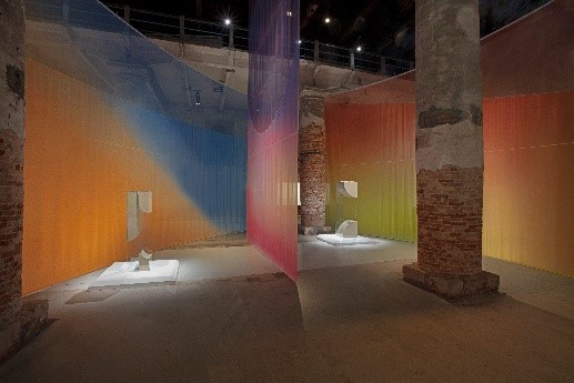 Art exhibition exhibit with translucent, blue, red, orange, red and green fabrics hanging vertically from the ceiling. The fabrics surround white sculptures, while the venue has brick columns and atmospheric lighting.