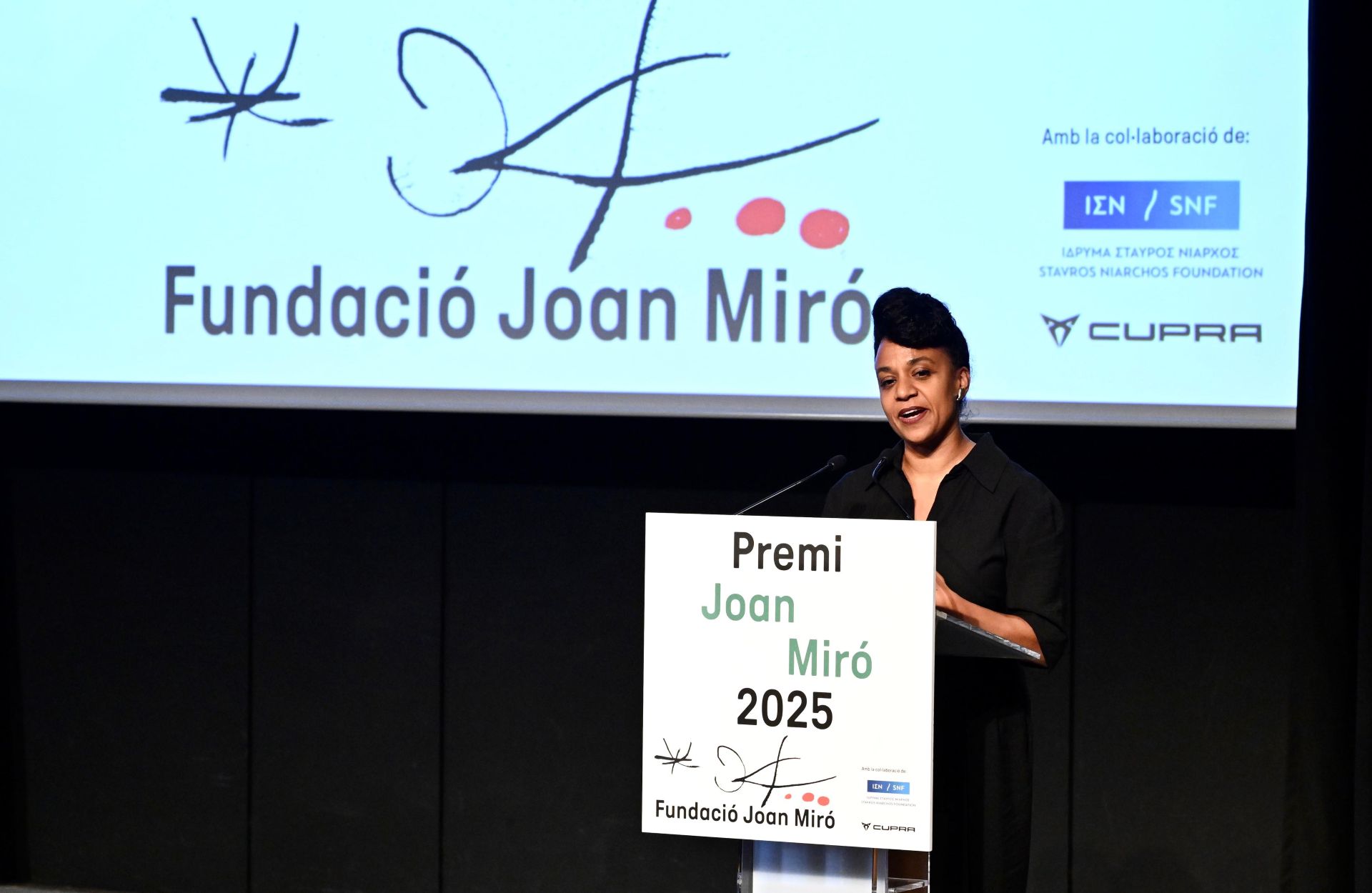A woman stands at a podium during the 2025 Joan Miró Prize event, with a screen behind her dispaying Fundació Joan Miró signage and the logos of the Stavros Niarchos Foundation and Cupra.