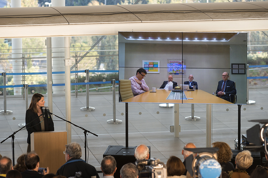 Μια φωτογραφία που δείχνει μια γυναίκα στο βήμα πάνω στη σκηνή να απευθύνει τον λόγο στους τέσσερις άντρες που συμμετέχουν μέσω διαδικτυακής σύνδεσης και το κοινό τους παρακολουθεί 