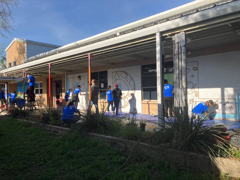 People in blue shirts working together to paint a building's exterior.