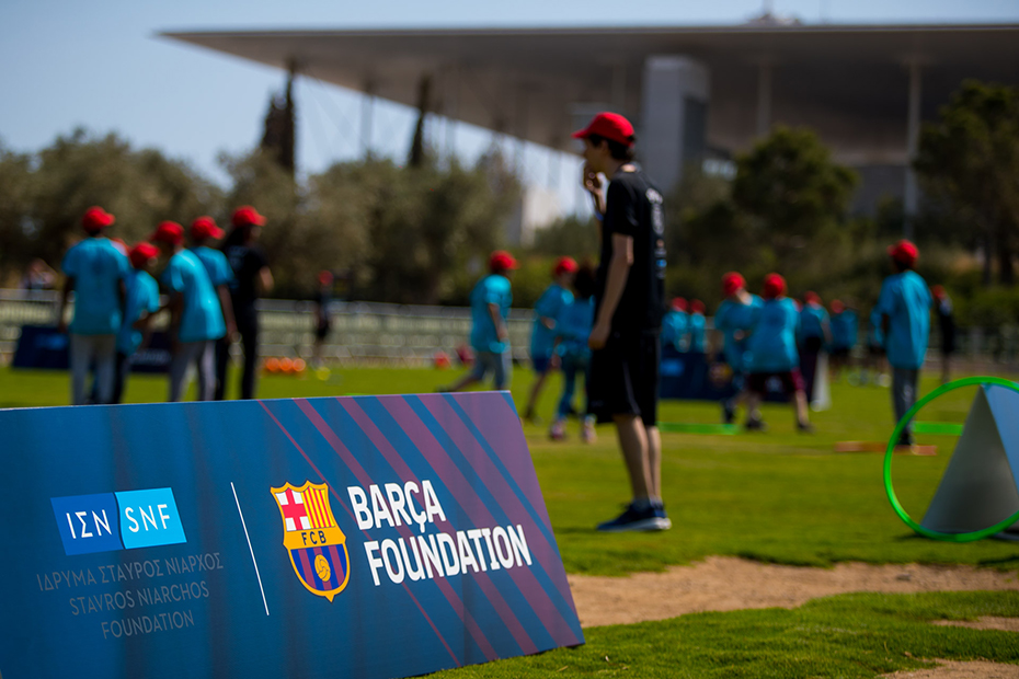 Παιδιά παίζουν σε ένα γήπεδο ποδοσφαίρου, με τα εμβλήματα του Barca Foundation να περιβάλλουν το γήπεδο