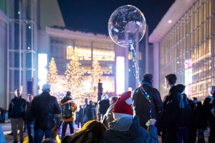 Φωτογραφία που δείχνει ένα παιδάκι με χριστουγεννιάτικο σκούφο να κρατάει ένα μπαλόνι και να παρακολουθεί με τον υπόλοιπο κόσμο τα φωτισμένα δέντρα που υπάρχουν στον χώρο ανάμεσα από δύο μεγάλα γυάλινα κτήρια 