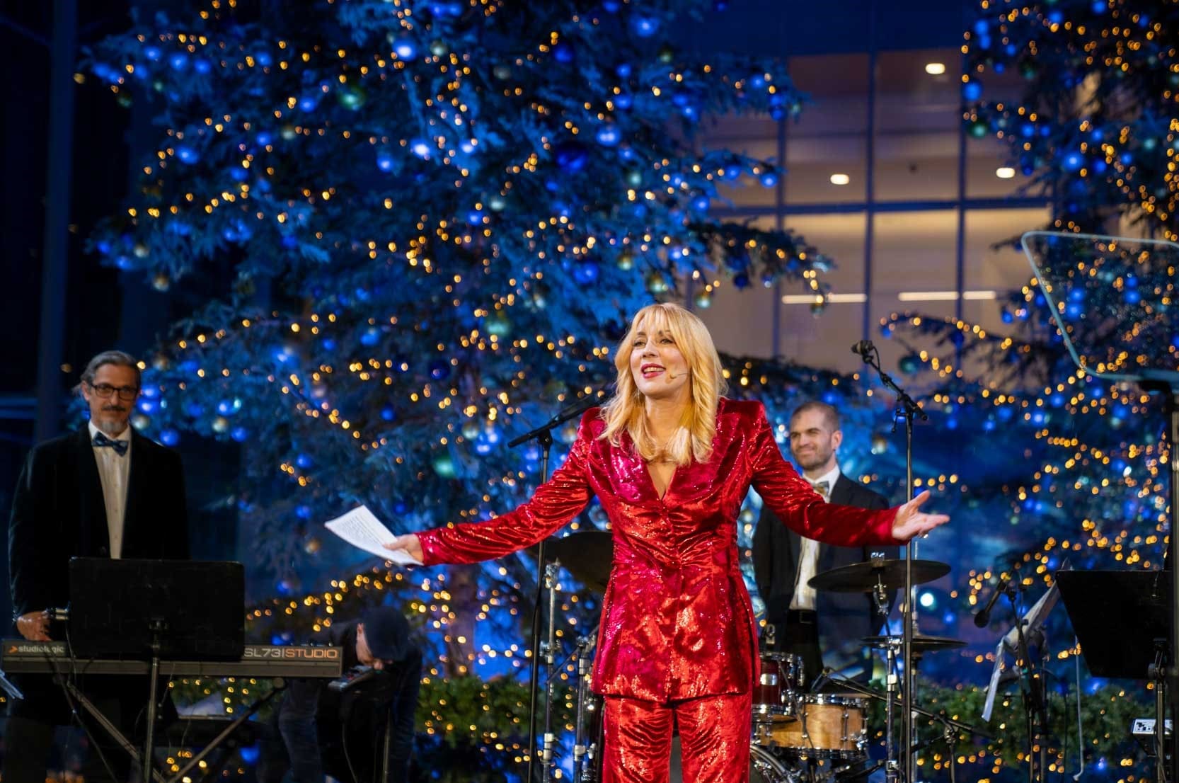 Η παρουσιάστρια με ένα λαμπερό κόκκινο κοστούμι εμφανίζεται σε μια σκηνή μπροστά από μεγάλα χριστουγεννιάτικα δέντρα.