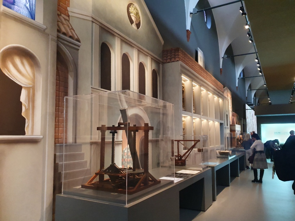 Φωτογραφία που δείχνει μια σειρά από ξύλινα εκθέματα μέσα σε διαφανή κουτιά τα οποία έχουν ως φόντο έναν αρχαιοελληνικό ναό 