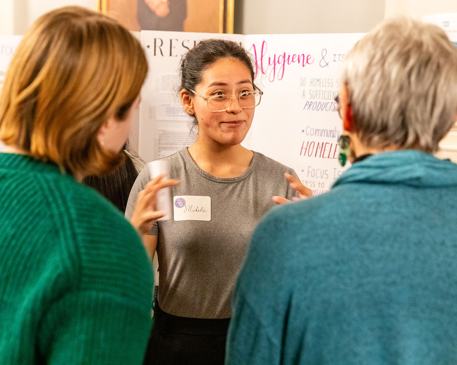 A high school student in front of a presentation posterboard speaks to two adult listeners.