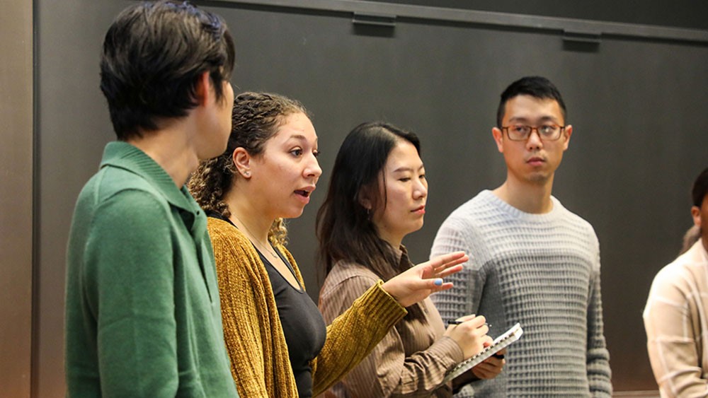 Μια ομάδα φοιτητών στέκονται όρθιοι, συζητούν και κρατάνε σημειώσεις 