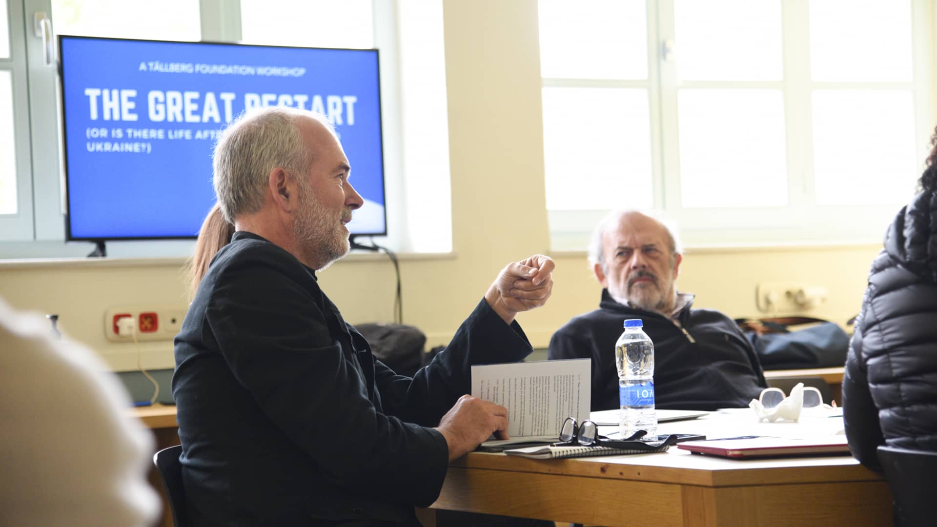 Ένας άντρας κάθεται στο τραπέζι και απευθύνει το λόγο σε άλλα μέλη του τραπεζιού 