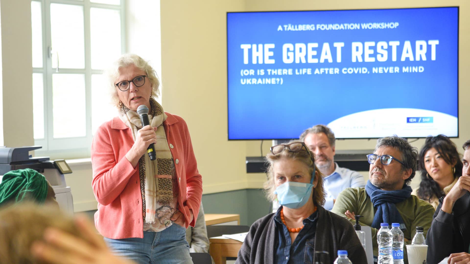 Μια γυναίκα στέκεται όρθια και μιλάει στο κοινό κρατώντας ένα μικρόφωνο και πίσω της υπάρχει μια οθόνη που γράφει 