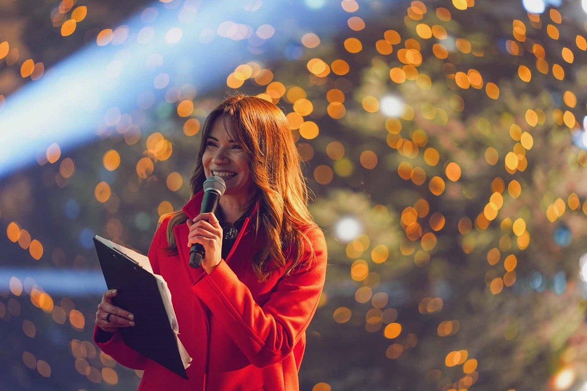 Μια γυναίκα κρτάει ένα ντοσιέ και ένα μικρόφωνο στα χέρια της και χαμογελάει