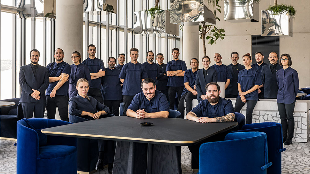 Μια ομάδα ανθρώπων ομοιόμορφα ντυμένη στέκεται όρθια για μια φωτογραφία, ενώ τρεις από αυτούς κάθονται στο τραπέζι μπροστά τους 