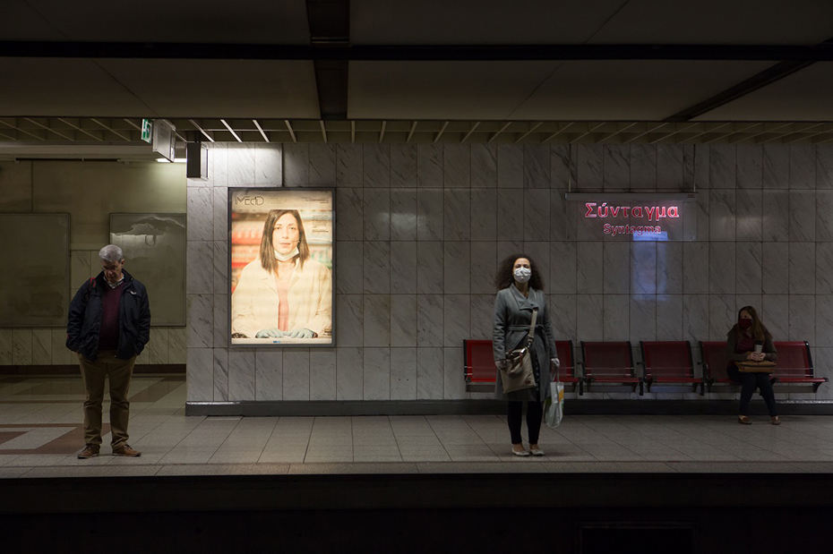 Στον σταθμό του μετρό Σύνταγμα υπάρχει ένας άντρας και μια γυναίκα που στέκονται όρθιοι, μια γυναίκα που κάθεται και μια οθόνη στην οποία προβάλεται μια γυανίκα και το λογότυπου του iMEdD