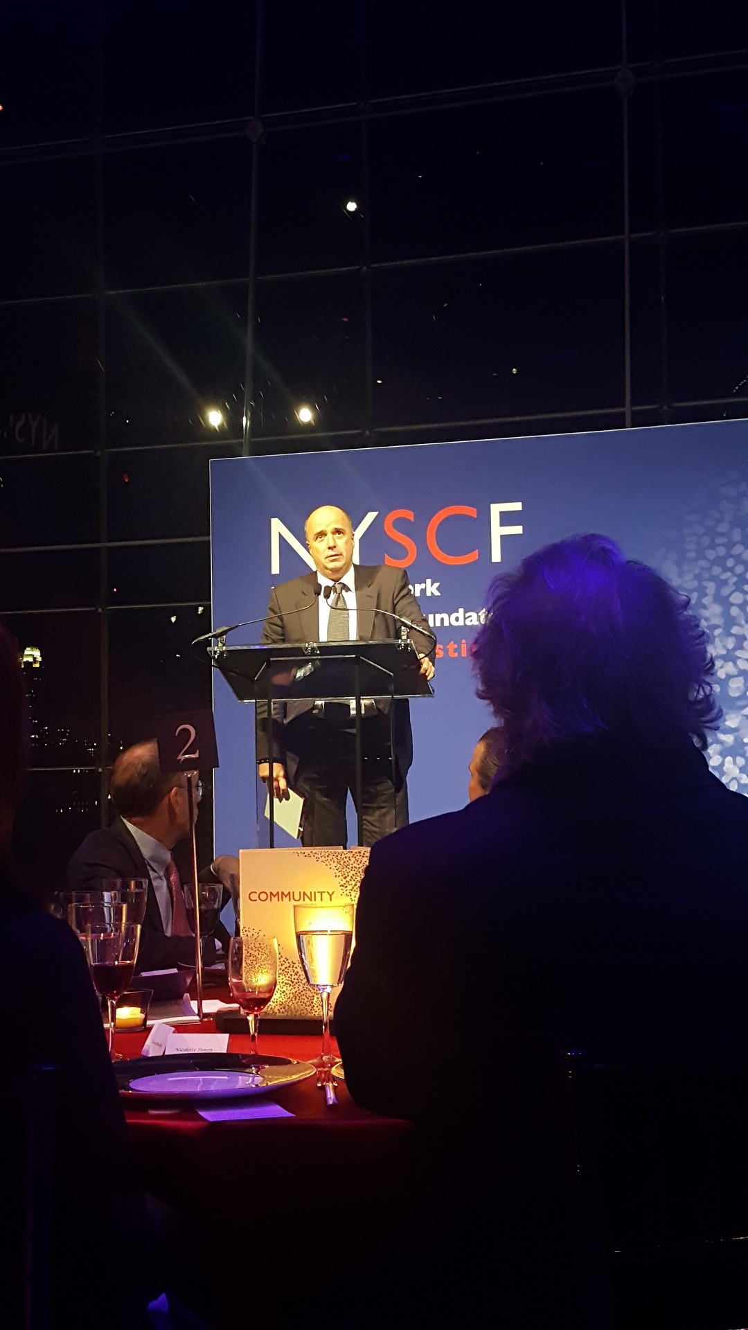Ο Αντρέας Δρακόπουλος, Πρόεδρος του Ιδρύματος Σταύρος Νιάρχος, μιλάει στο βήμα του NYSCF και ο κόσμος από κάτω κάθεται σε τραπέζια και τον παρακολουθεί 