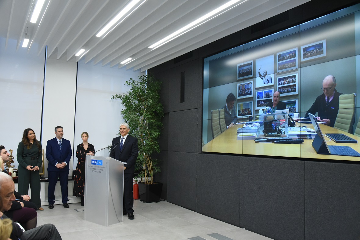 Φωτογραφία τραβηγμένη από το πλαι η οποία δείχνει κάποιους να κάθονται σε καρέκλες, άλλους να στέκονται όρθιοι, ένας άντρας να στέκεται στο βήμα και όλοι παρακολουθούν τη ζωντανή σύνδεση με τον Αντρέα Δρακόπουλο, Πρόεδρο του Ιδρύματος Σταύρος Νιάρχος 