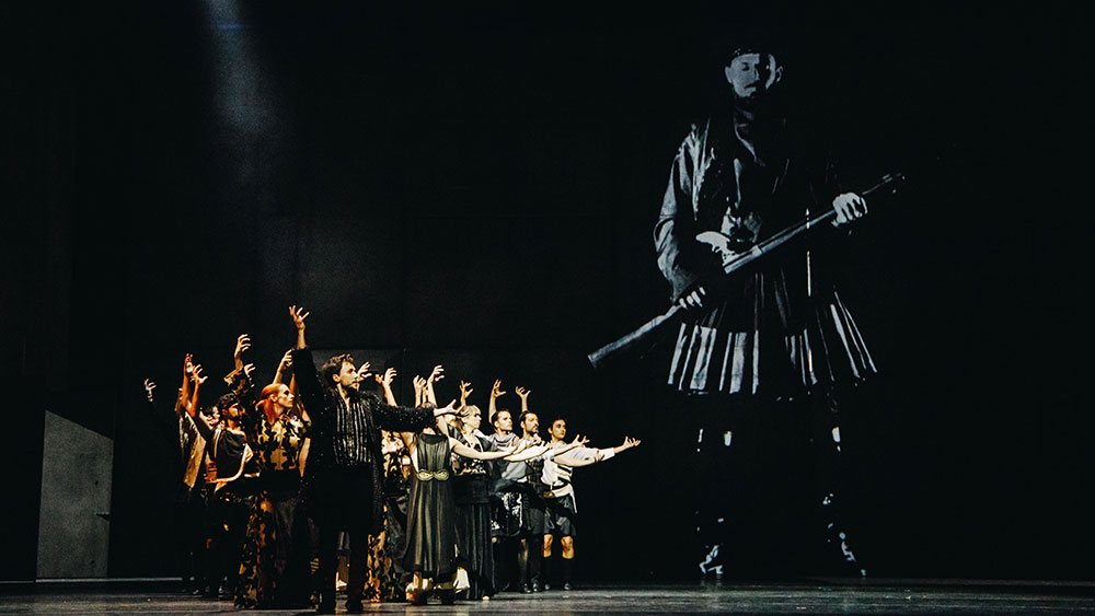 Πάνω σε μια σκηνή μια ομάδα ηθοποιών ντυμένοι με ελληνικές στολές πολέμου στέκονται και δείχνουν με τα χέρια τους μια φωτογρφία ενός άντρα από τον πόλεμο που στέκεται με το όπλο του 