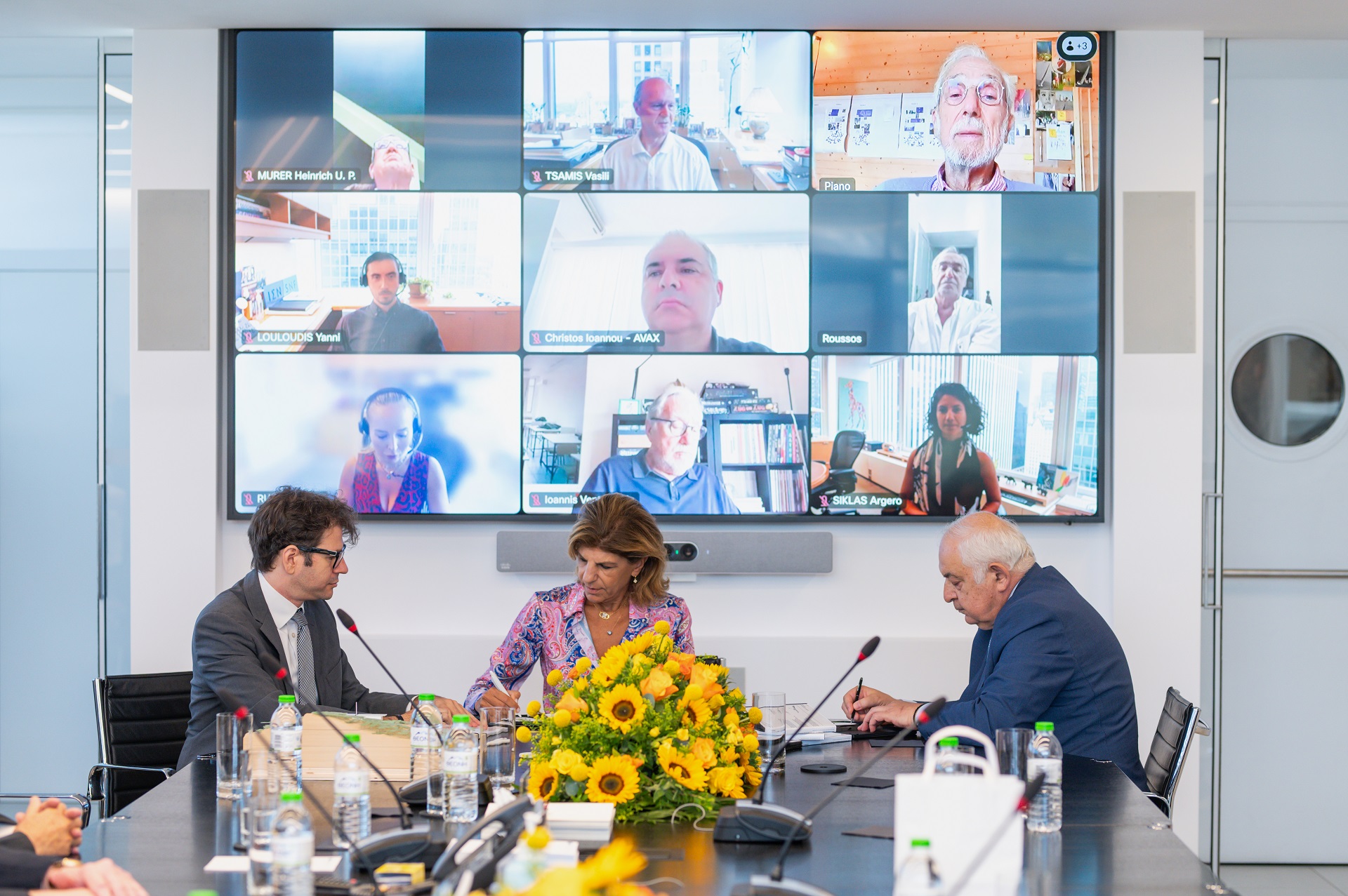 Δύο άντρες και μια γυναίκα μπροστά από ζωντανή σύνδεση με άλλα μέλη του Ιδρύματος Σταύρος Νιάρχος, υπογράφουν για την κατασκευή των τριών νέων νοσκομείων Ίδρυμα Σταύρος Νιάρχος 