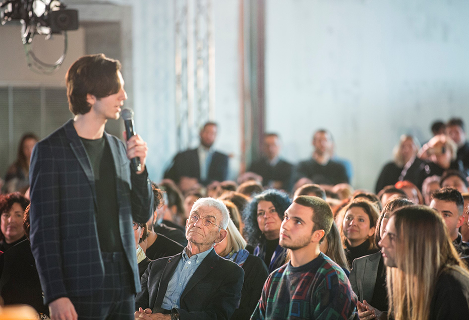 Ένας νεαρός άντρας από το κοινό στέκεται όρθιος και κρτάει ένα μικρόφωνο καθώς εκφράζει τον προβληματισμό του 