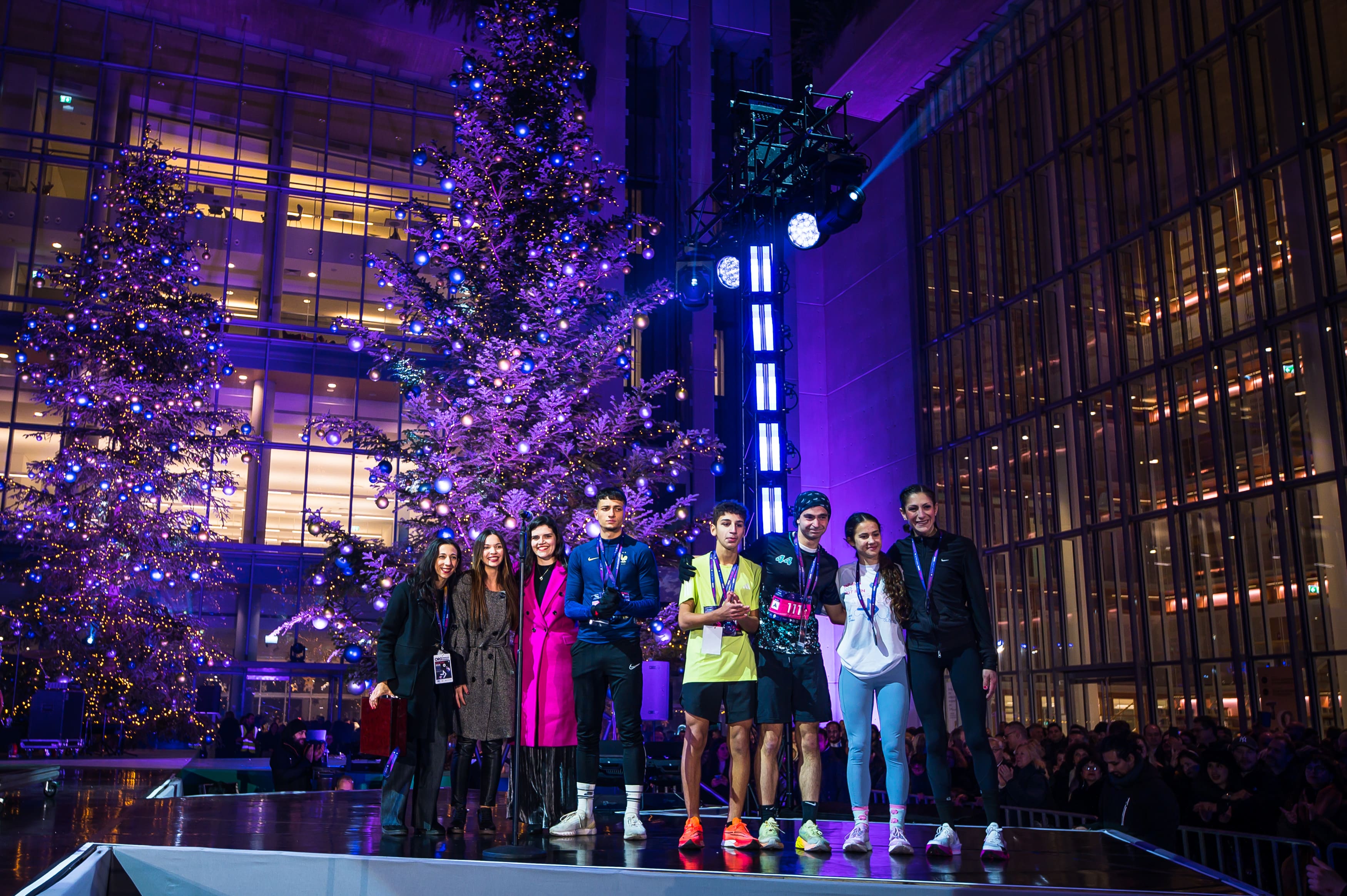 8 άτομα που στέκονται στη σειρά πάνω στη σκηνή του ΚΠΙΣΝ, μπροστά από χριστουγεννιάτικα δέντρα.