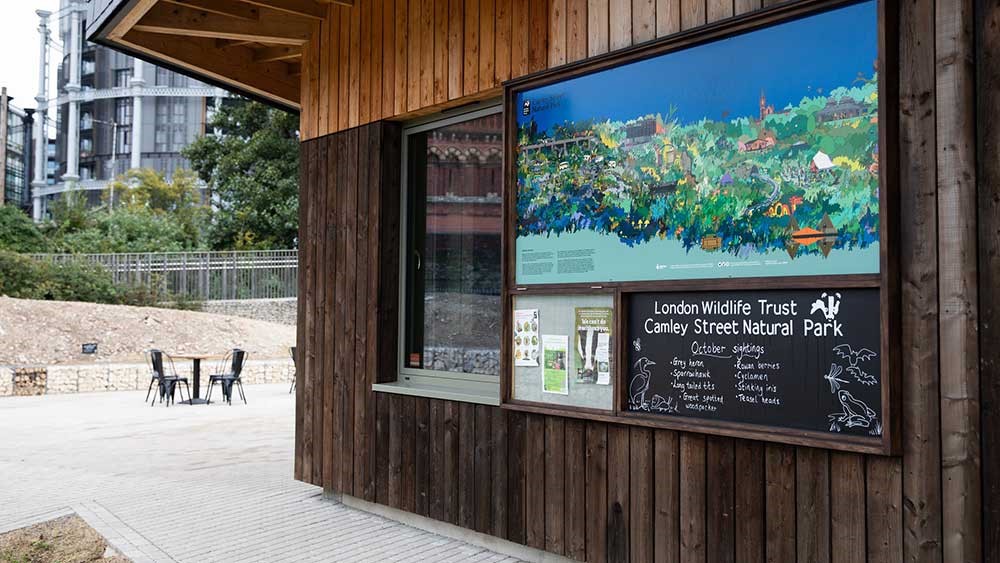 Φωτογραφία από το ξύλινο κιόσκι στον εξωτερικό χώρο του Κέντρου Επισκεπτών και Εκπαίδευσης Ίδρυμα Σταύρος Νιάρχος στο οποίο υπάρχει ένας πίνακας με ζωγραφιά και αφίσες και γράφει London Wildlife Trust Camley Street Natural Park 