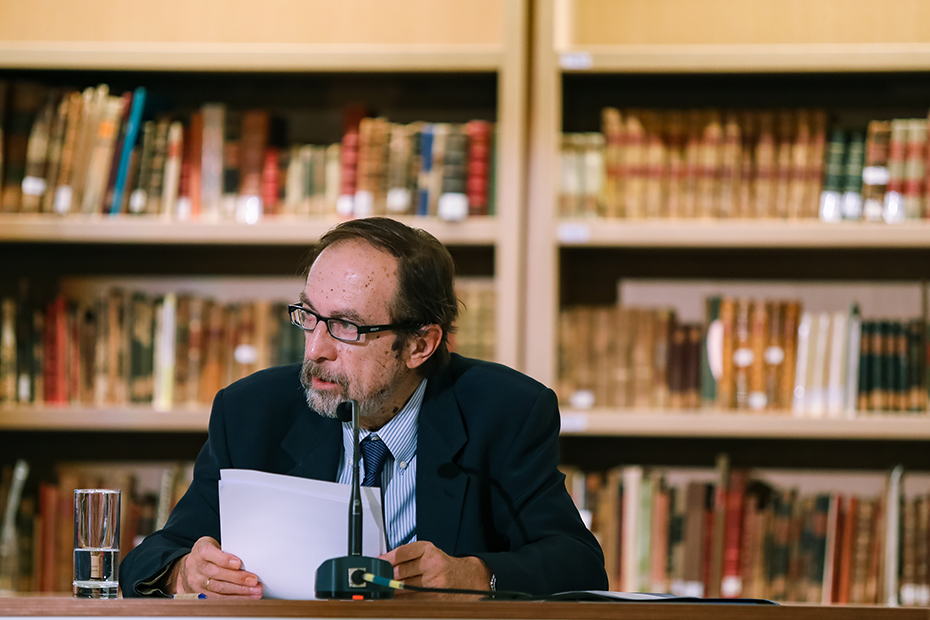 Ένας άντρας κάθεται στη σκηνή και μιλάει στο μικρόφωνο κρατώντας χαρτιά στα χέρια του 