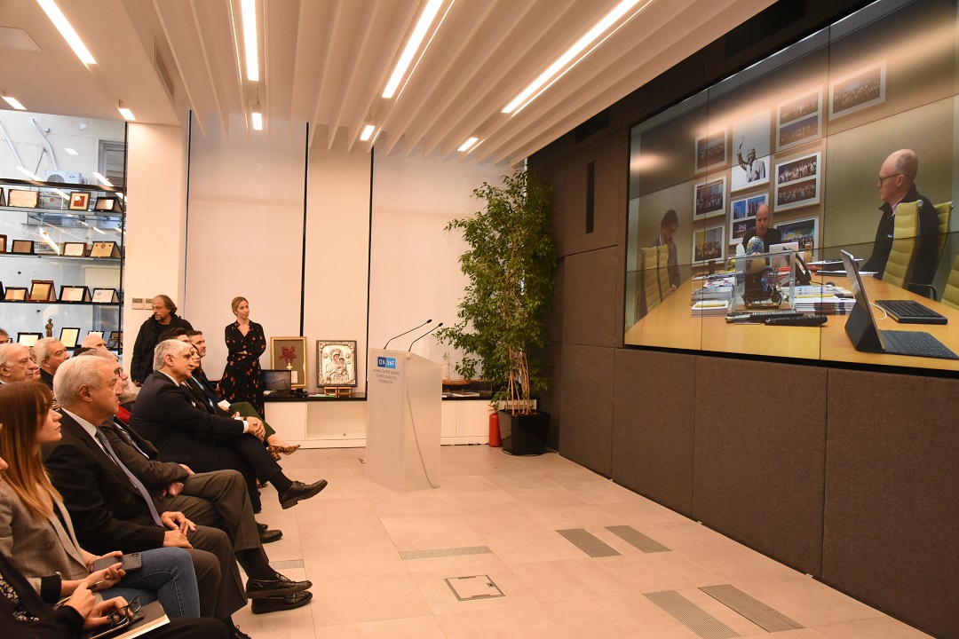 Άνθρωποι κάθονται σε καρέκλες και παρακολουθούν τη ζωντανή σύνδεση με τον Αντρέα Δρακόπουλο, Πρόεδρο του Ιδρύματος Σταύρος Νιάρχος 