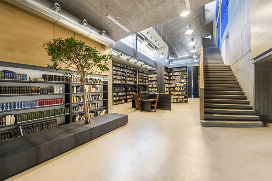 Φωτογραφία που δείχνει τον εσωτερικό χώρο μιας βιβλιοθήκης στον οποίο υπάρχουν ράφια γεμάτα βιβλία, τρπέζια με καρέκλες, μια σκάλα που οδηγεί σε άλλον όροφο και ένα μεγάλο παγκάκι με ένα δέντρο τοποθετημένο πάνω του