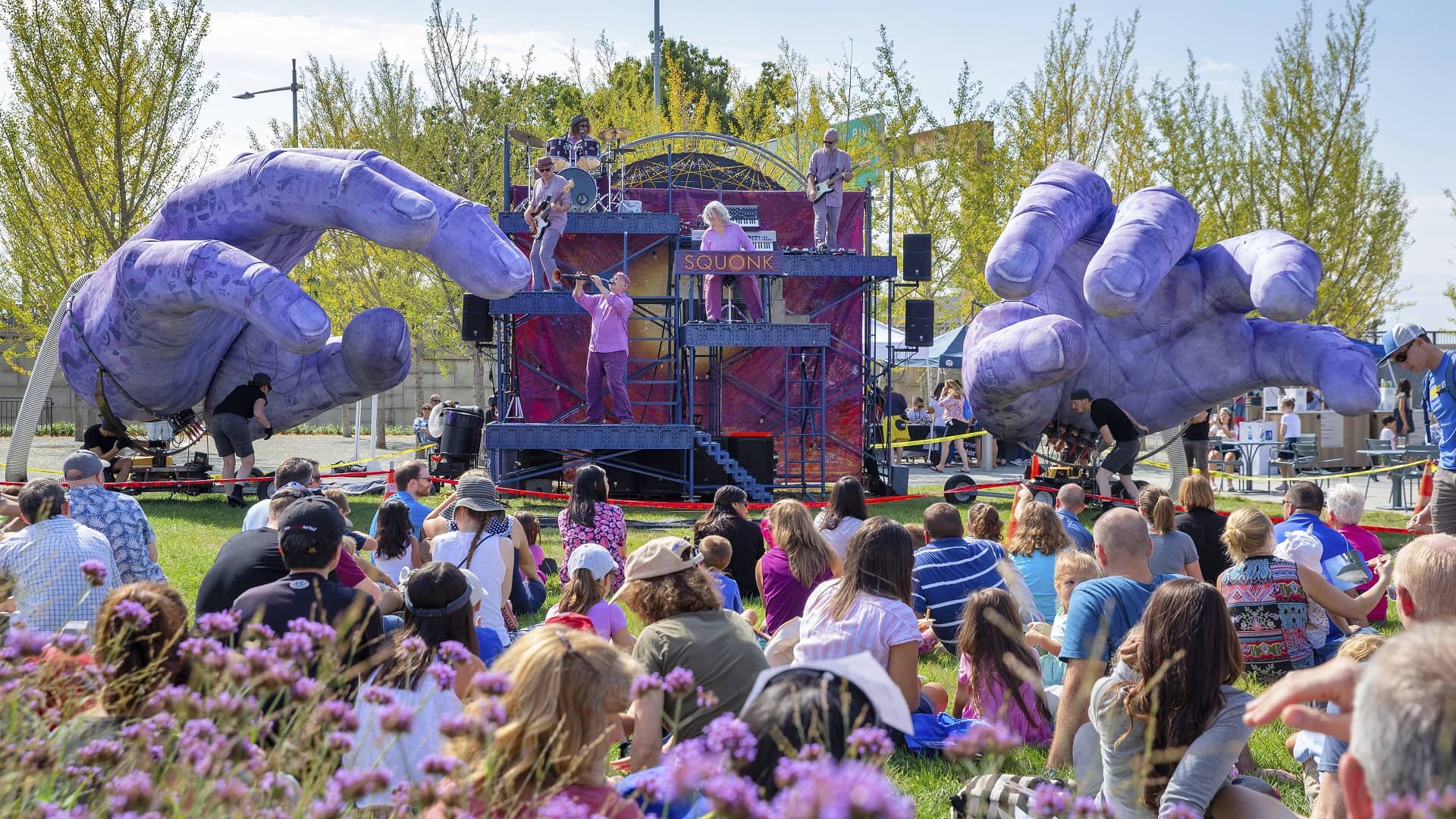 Πλήθος ανθρώπων κάθονται στο γρασίδι και παρακολουθούν μια συναυλία όπου έχει ως διακόσμηση η σκηνή δύο μεγάλα μωβ χέρια 