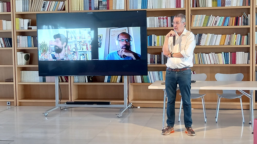 Ένας άντρας στέκεται όρθιος και μιλάει κρατώντας ένα μικρόφωνο και δίπλα του έχει μια μεγάλη οθόνη που δείχνει τη διαδικτυακή σύνδεση με δύο άλλους άντρες 