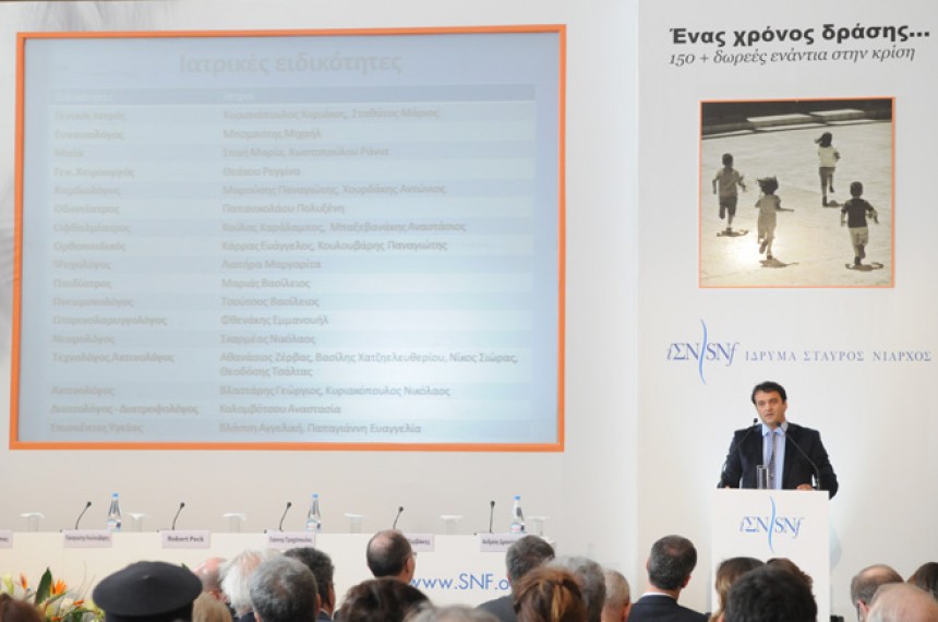 Ένας άνδρας μιλάει στη σκηνή στη συνέντευξη Τύπου του Ιδρύματος Σταύρος Νιάρχος δίπλα σε μια μεγάλη οθόνη που απαριθμεί ονόματα γιατρών και τις ιατρικές τους ειδικότητες.