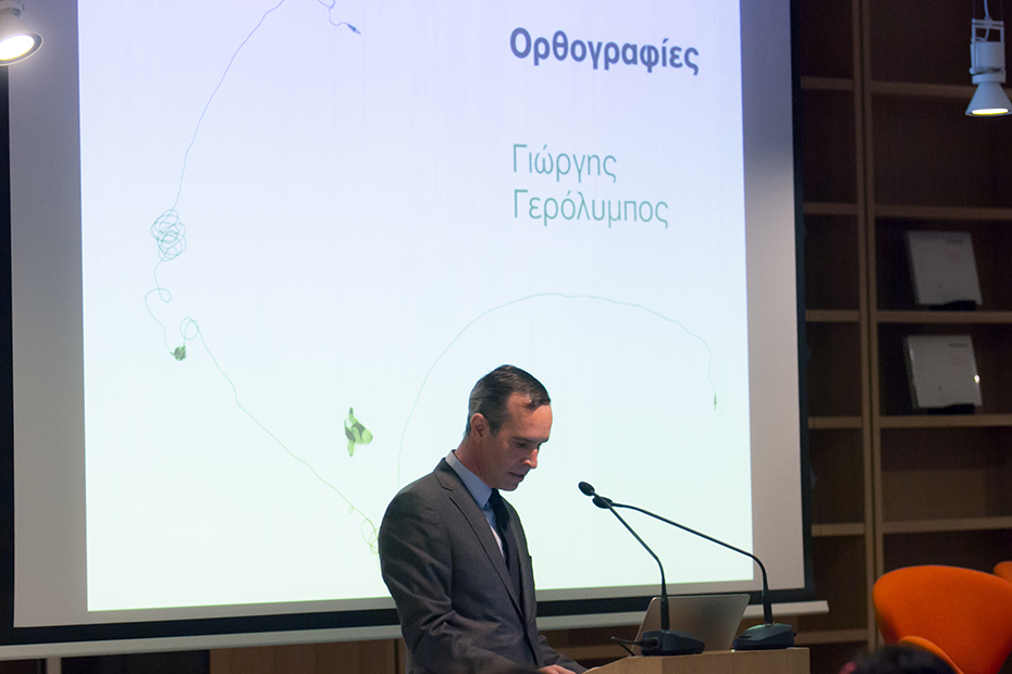 Ένας άντρας στη σκηνή στο βήμα ετοιάζεται να βγάλει λόγο ενώ πίσω του προβάλλεται στον προτζέκτορα μια εικόνα που γράφει Ορθογραφίες Γιώργης Γερόλυμπος