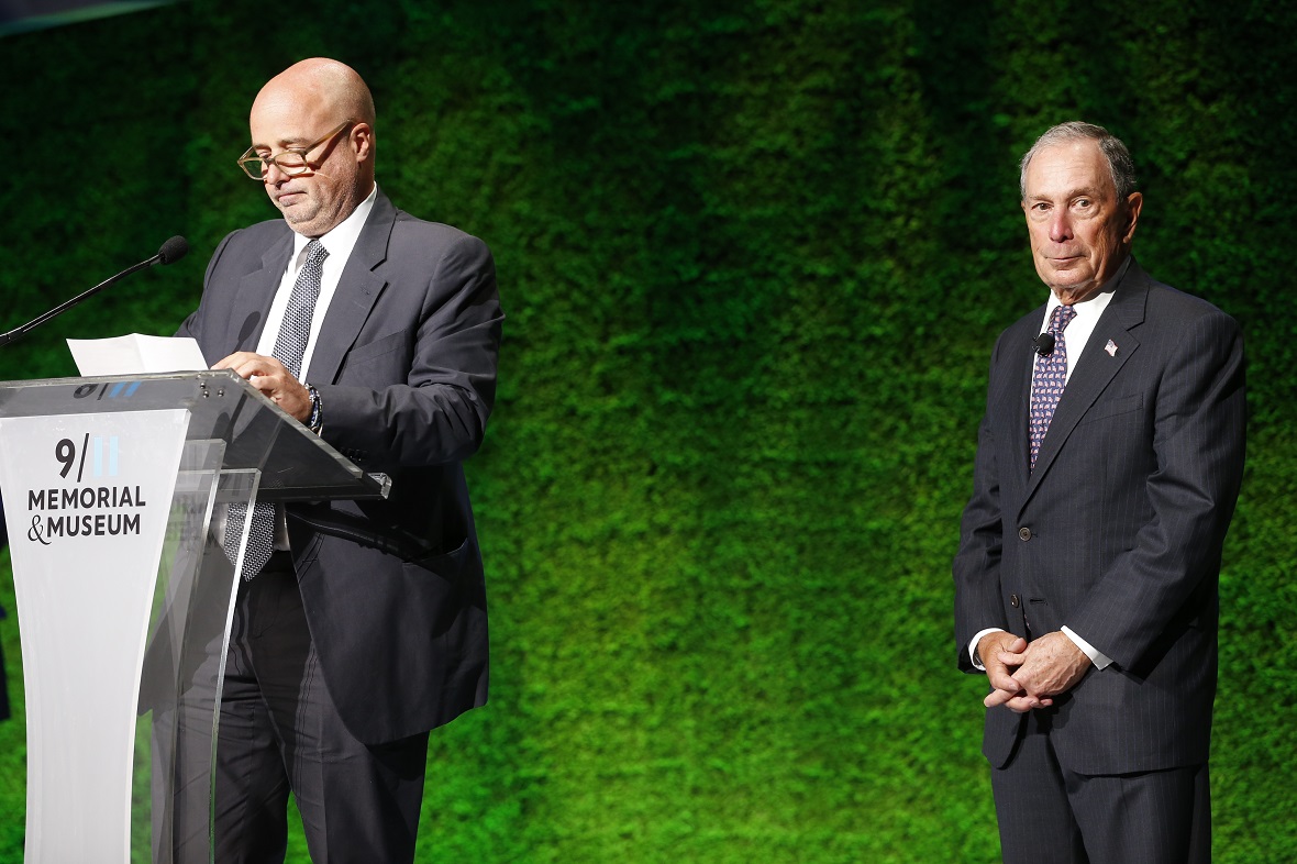 Andreas Dracopoulos, President of the Stavros Niarchos Foundation, on the podium of the 9 I II Memorial & Museum, looks at his papers, while a man stands behind him and watches him 