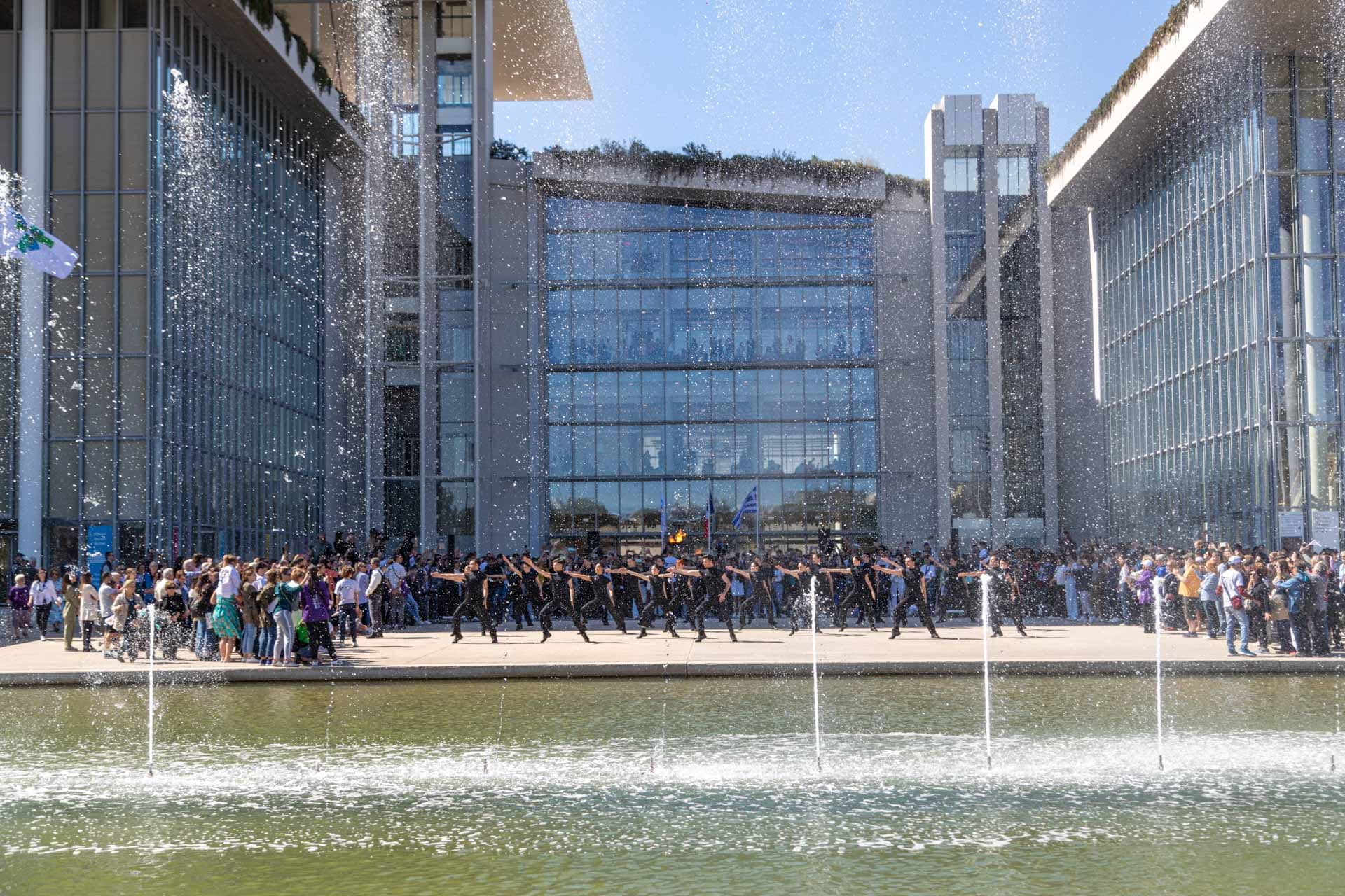 Μια χορευτική παράσταση, περιτριγυρισμένη από πλήθος με φόντο το Κέντρο Πολιτισμού Ίδρυμα Σταύρος Νιάρχος, φαίνεται μέσα από συντριβάνια σε πρώτο πλάνο.