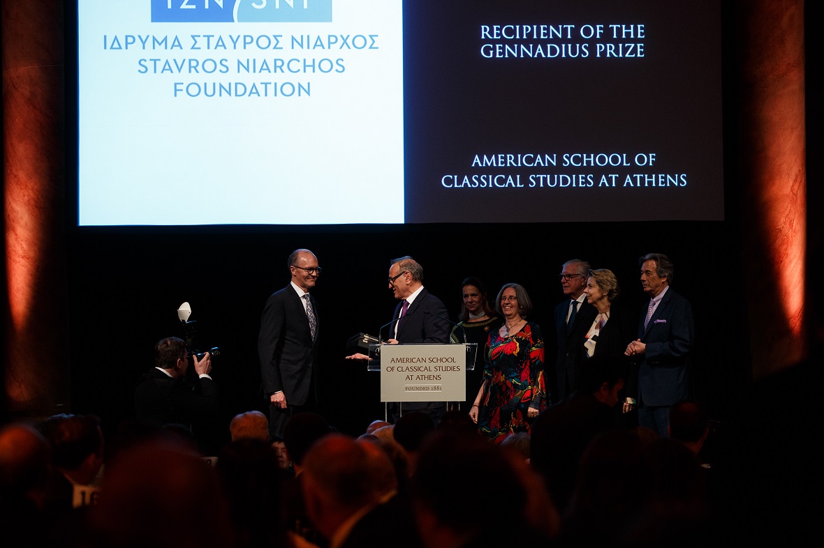 Members of the Stavros Niarchos Foundation stand on stage to receive the Gennadius Prize in front of a screen that writes 