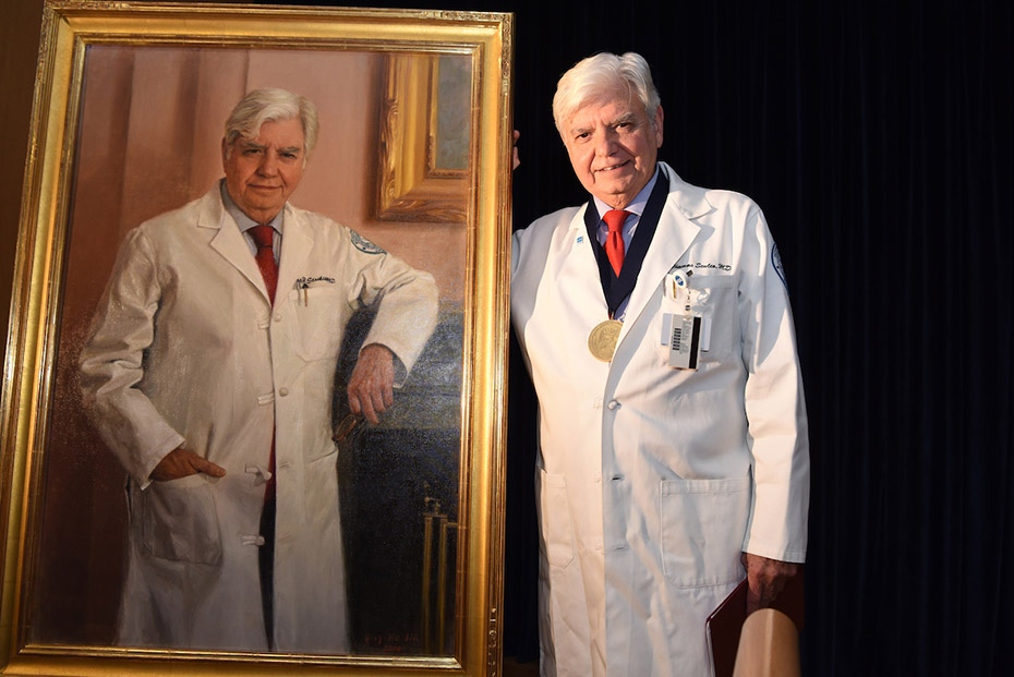 Ένας άντρας με ιατρική στολή φοράει ένα μετάλιο και στέκεται δίπλα στο πορτρέτο του