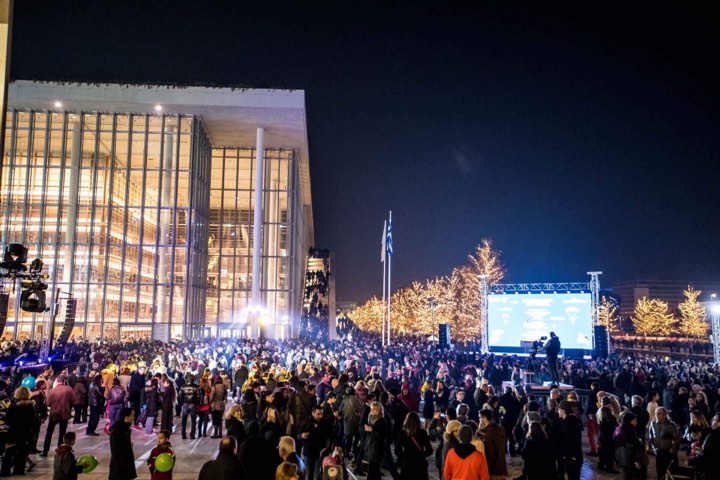 Πλήθος ανθρώπων παρακολουθεί τη σκηνή που έχει στηθεί δίπλα από ένα μεγάλο γυάλινο κτήριο 