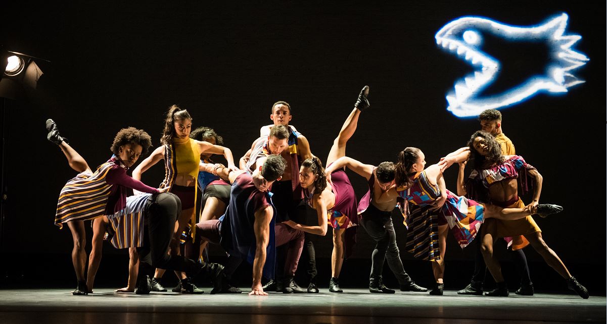 Μια ομάδα χορευτών πάνω στη σκηνή ποζάρουν κάνοντας φιγούρες τέτοιες ώστε να φαίνεται ότι είναι μπλεγμένοι μεταξύ τους