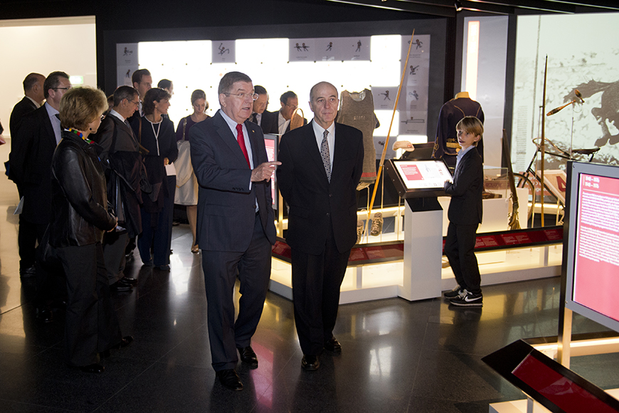 Δυο άντρες περπατούν και συζητούν σε μια έκθεση και ο κόσμος γύρω τους κοιτάει τα εκθέματα