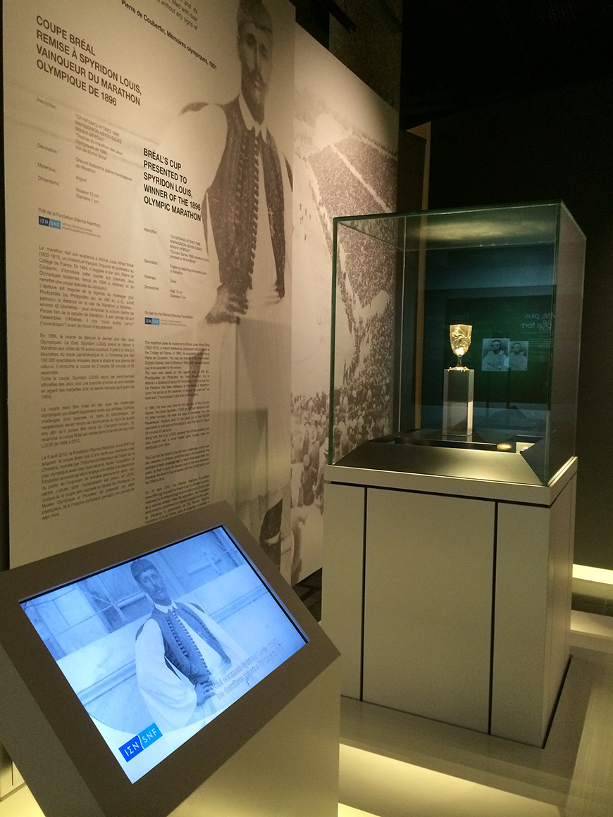 Το κύπελλο του Σπύρου Λούη κλεισμένο σε γυαλί δίπλα σε έναν τοίχο με πληροφορίες για τη σημασία του