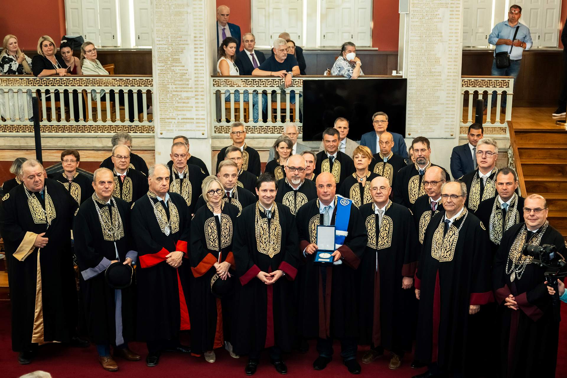 Ο τιμώμενος Πρόεδρος του ΙΣΝ, Ανδρέας Δρακόπουλος, κρατά τιμητική πλακέτα, και στέκεται όρθιος ανάμεσα σε μέλη της Συγκλήτου και της ακαδημαϊκής κοινότητας του ΕΚΠΑ. Όλοι τους είναι ντυμένοι με τηβέννους. 