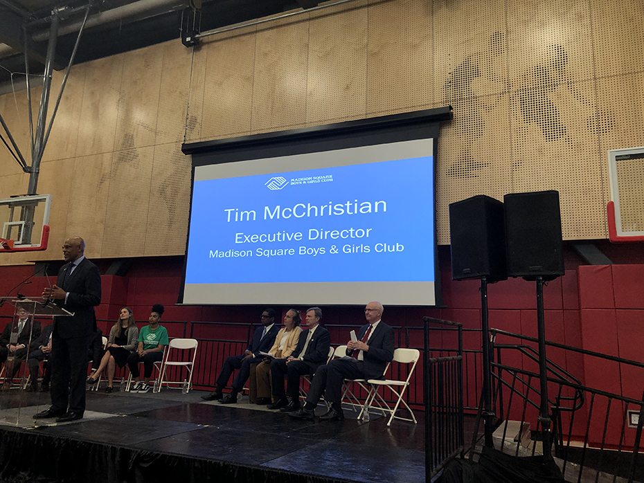 Σε μια σκηνή υπάρχουν κάποιοι άντρες οι οποίοι κάθονται και ένας άντρας είναι στο βήμα και βγάζει λόγο ενώ πίσω τους προβάλλεται σε μια οθόνη μια φωτογραφία που γράφει Tim McChristian Executive Director Madison Square Boys & Girls Club